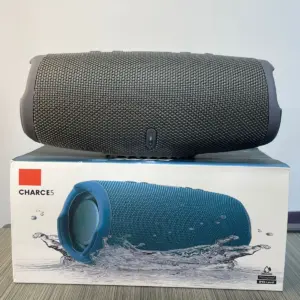 Parlante Bluetooth JBL Charge 5 gris oscuro sobre su caja original, resaltando su diseño cilíndrico y resistencia al agua.