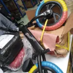 Bicicleta de equilibrio infantil sin pedales, marco negro y llantas multicolores azul, amarillo y rojo, junto a su empaque en mesa blanca.