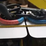 Bicicleta de equilibrio infantil plegada con llantas bicolor rojo y azul, marco negro y cinta métrica para referencia de tamaño sobre mesa blanca.