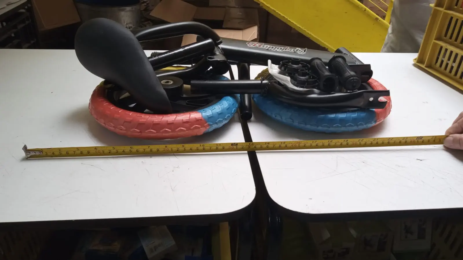 Bicicleta de equilibrio infantil plegada con llantas bicolor rojo y azul, marco negro y cinta métrica para referencia de tamaño sobre mesa blanca.