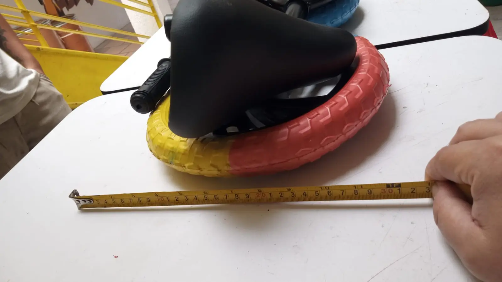 Bicicleta de equilibrio con asiento negro y llanta bicolor amarillo y rojo, mostrando dimensiones con cinta métrica sobre mesa blanca.