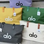 Cinco bolsos de lona Alo en colores morado, amarillo, verde, gris y blanco, con asas de cuero y el logotipo impreso, exhibidos sobre escalones.