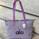 Bolso tote morado con textura tejida y logotipo alo negro, asas largas de cuero sintético y etiqueta colgante sobre escalones de piedra.