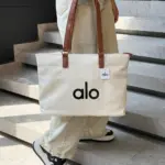 Bolso de lona blanco marfil de gran capacidad con asas de cuero marrón y la marca Alo en negro, sostenido por una persona en escaleras.