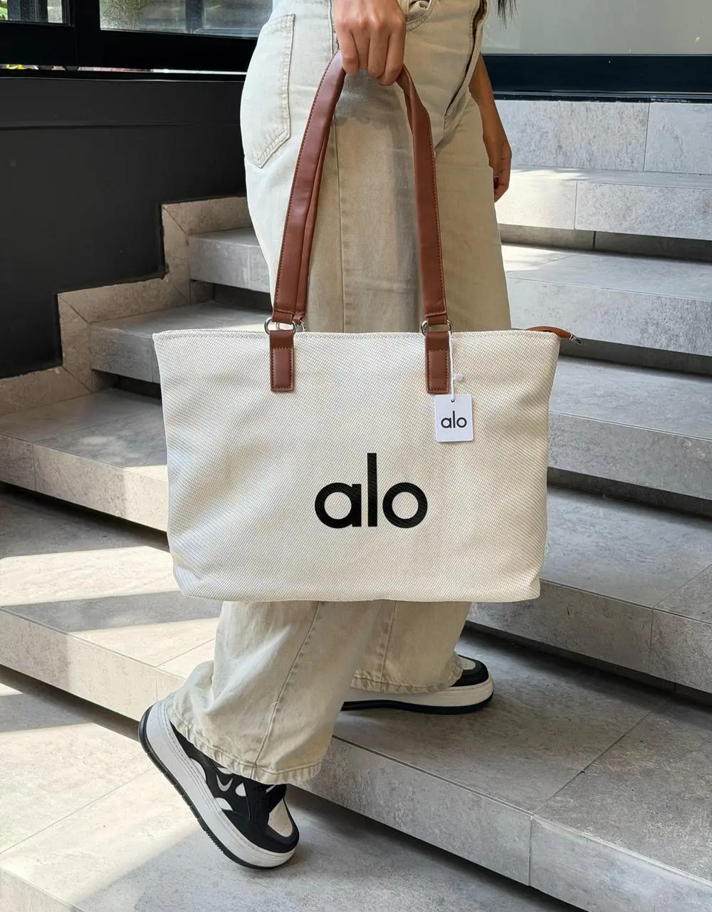 Bolso de lona blanco marfil de gran capacidad con asas de cuero marrón y la marca Alo en negro, sostenido por una persona en escaleras.