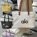 Bolso Alo beige de gran capacidad con asas marrones, rodeado de variantes en verde, amarillo, negro, lila y crema sobre escaleras.