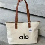 Bolso grande de lona beige con estampado alo negro y asas de cuero marrón, sostenido sobre escaleras de piedra.