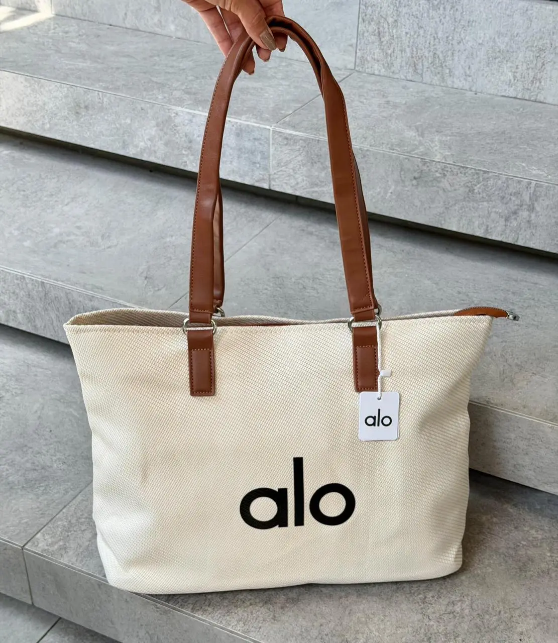 Bolso grande de lona beige con estampado alo negro y asas de cuero marrón, sostenido sobre escaleras de piedra.