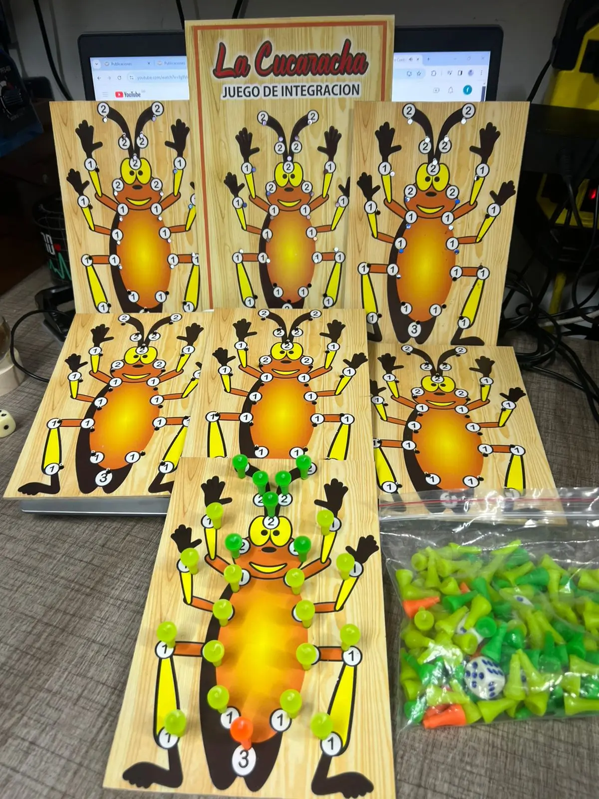 Tableros de madera del juego La Cucaracha con diseño de cucaracha, junto a una bolsa con fichas plásticas verdes y naranjas.