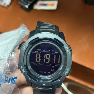 Reloj deportivo digital negro con pantalla LCD, carcasa texturizada y botones laterales, sostenido en mano sobre fondo de madera.