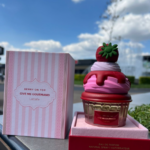 Perfume en envase con forma de cupcake de fresa con crema, 75 ml, junto a su caja rosa a rayas frente a una fuente y cielo nublado.