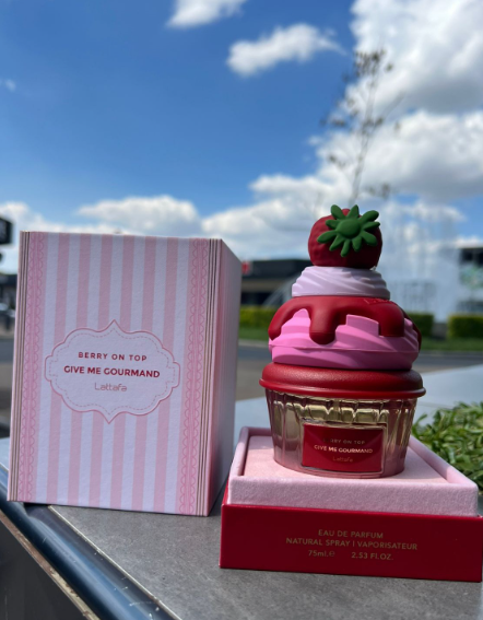 Perfume en envase con forma de cupcake de fresa con crema, 75 ml, junto a su caja rosa a rayas frente a una fuente y cielo nublado.