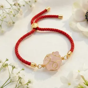 Pulsera artesanal roja con dije de cuarzo rosa en bruto envuelto en alambre dorado, sobre fondo blanco con flores.