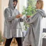 Pareja sonriendo en casa usando busos térmicos oversize grises de tela afelpada con capucha y bolsillo frontal, disfrutando de gran comodidad y abrigo.