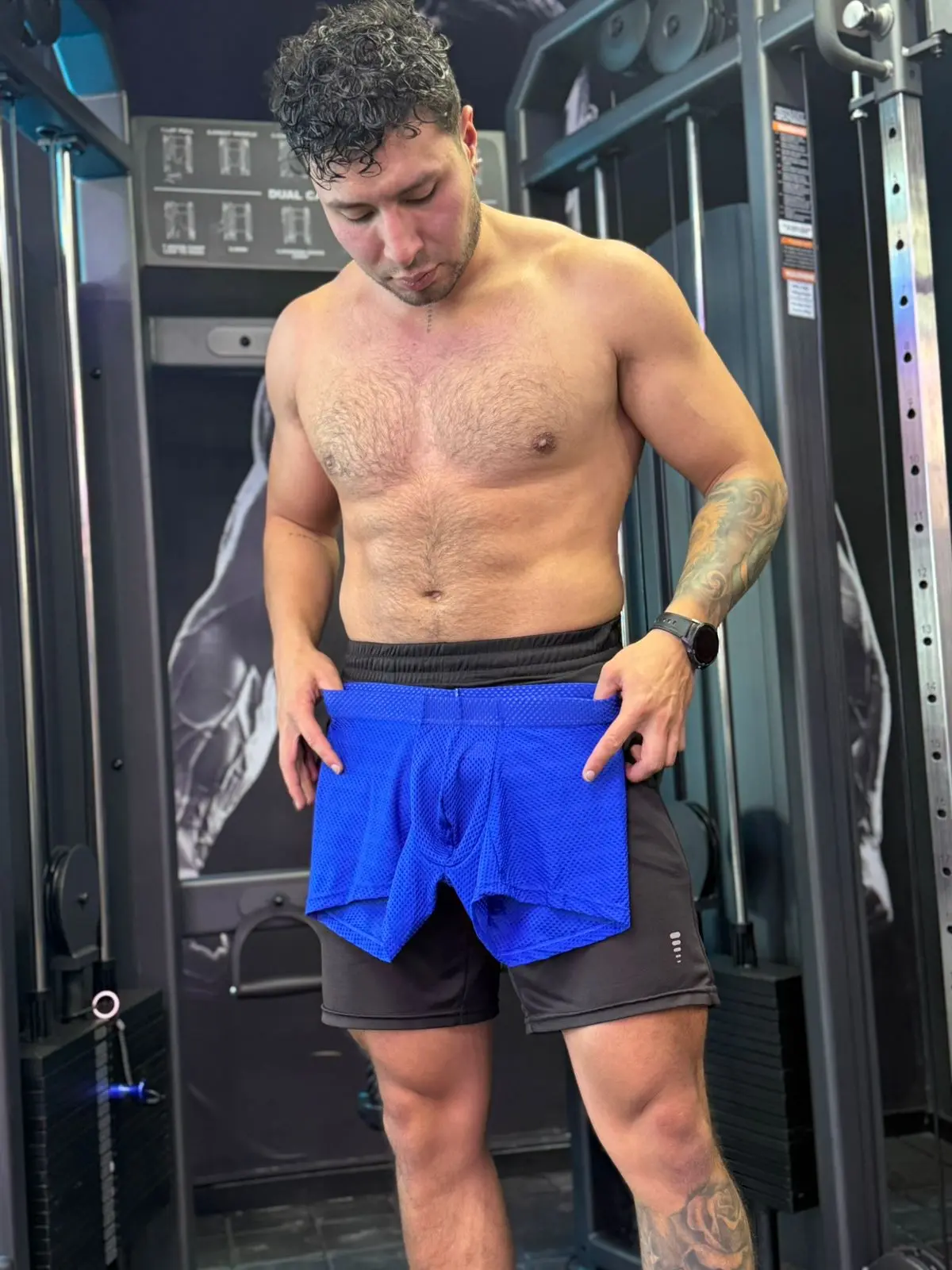 Hombre musculoso sosteniendo bóxer azul de tela licrada microperforada en el gimnasio, destacando su textura transpirable.