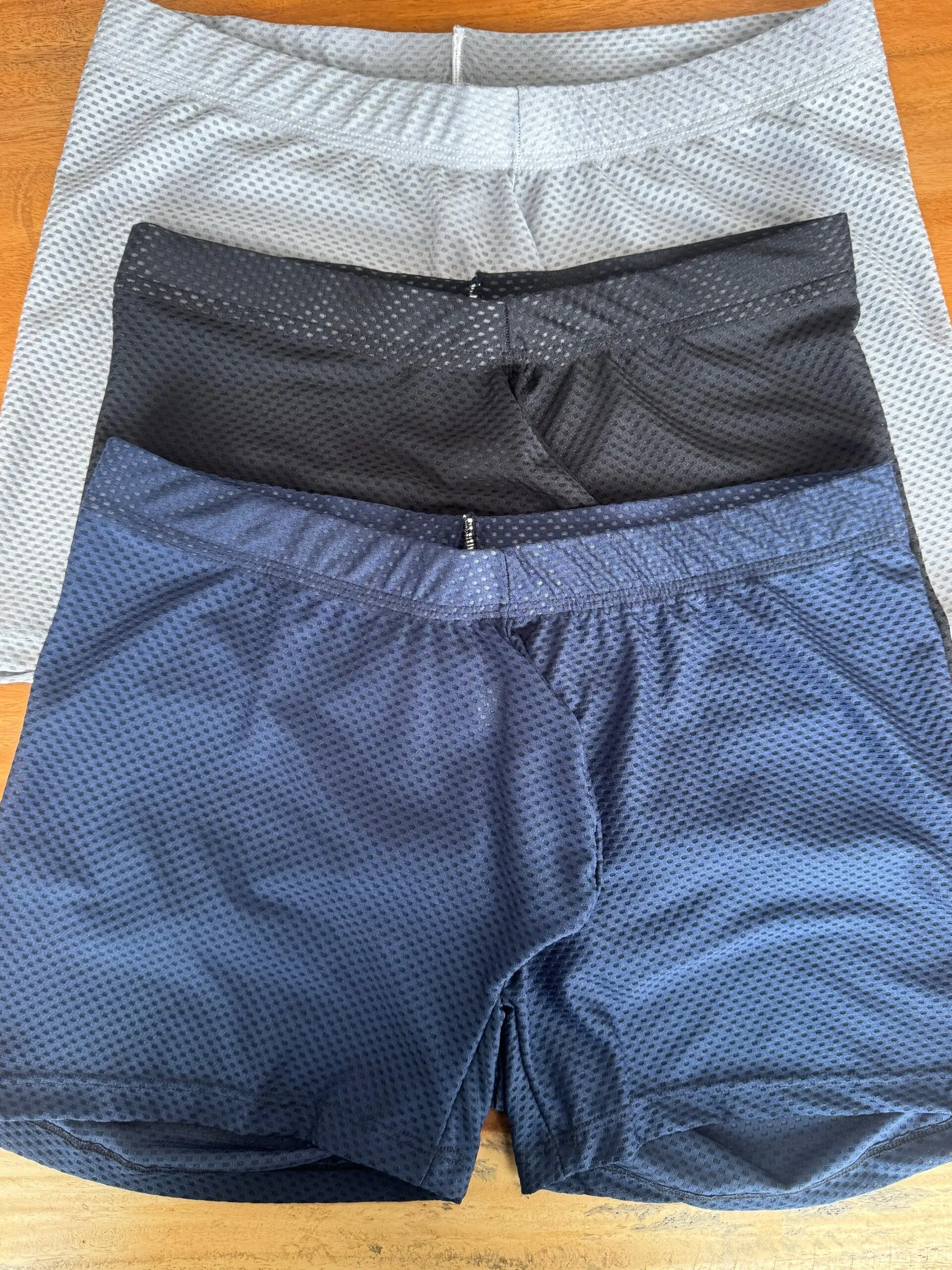 Tres bóxers transpirables de tela licrada con diseño microperforado en colores gris, negro y azul marino, apilados verticalmente sobre superficie de madera.