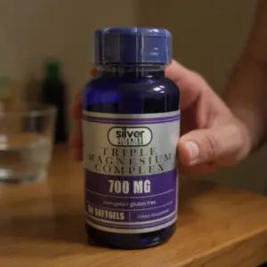 Frasco azul translúcido con tapa de seguridad, etiqueta plateada y morada que indica magnesio complejo 700 mg, presentación de 50 cápsulas blandas sobre mesa de madera.