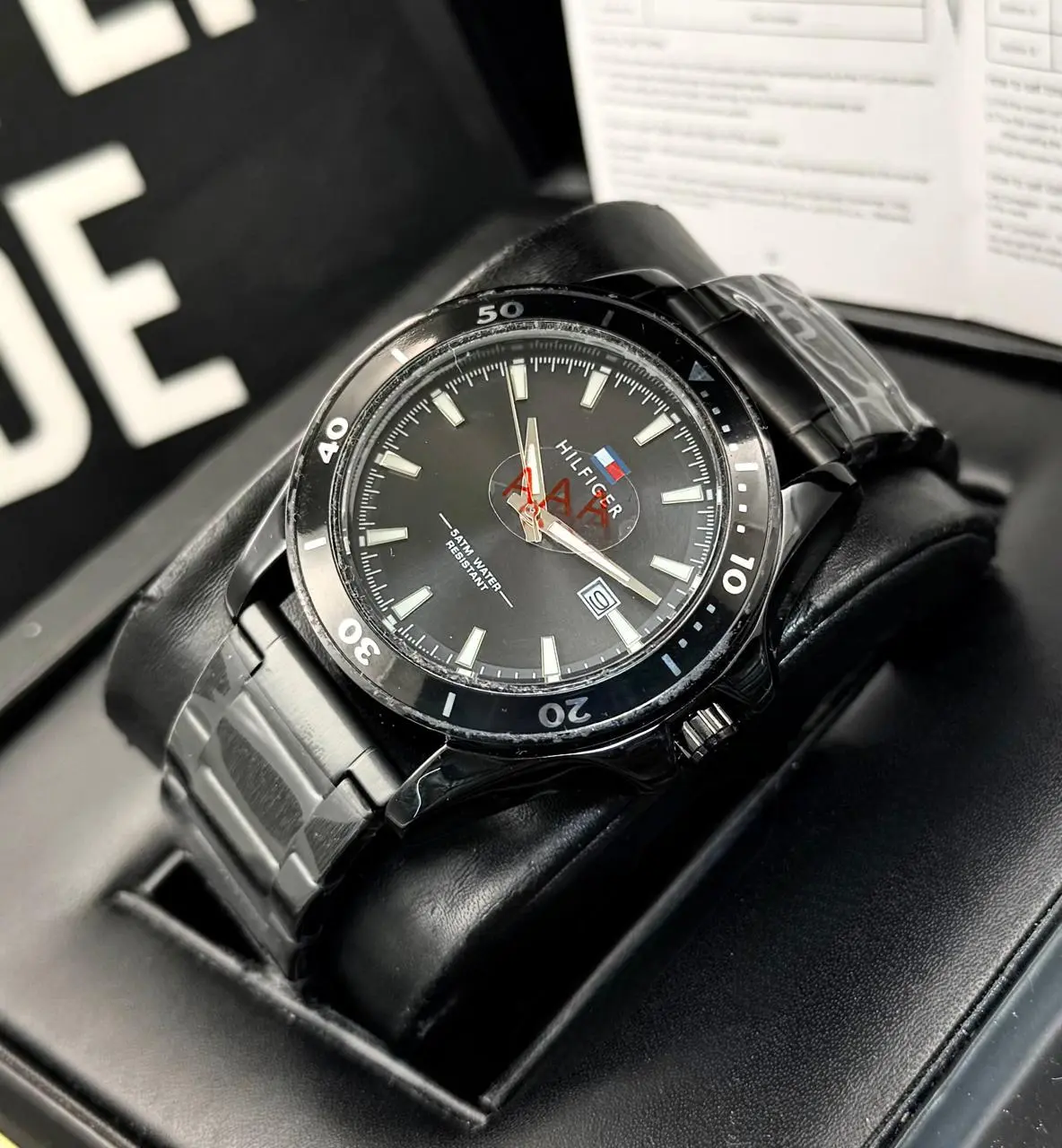 Reloj analógico Tommy Hilfiger para hombre en acero negro con esfera oscura, bisel giratorio, calendario y resistencia al agua, montado sobre cojín en su estuche.