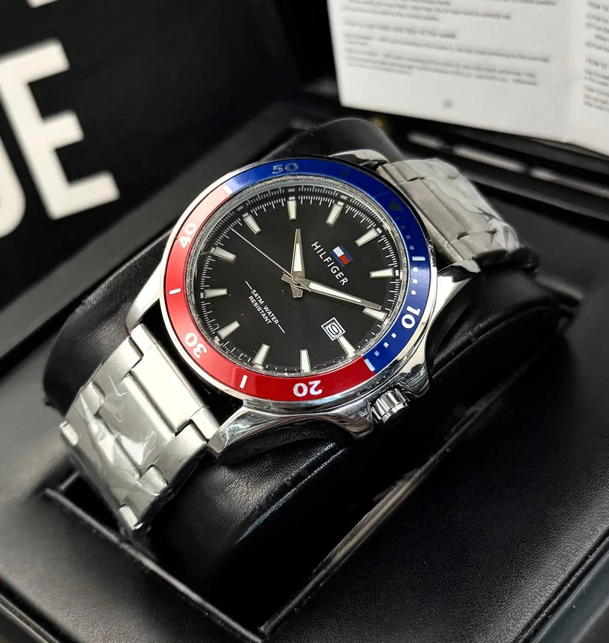 Reloj Tommy analógico de acero plateado con esfera negra, bisel giratorio rojo y azul, y calendario, sobre cojín negro.