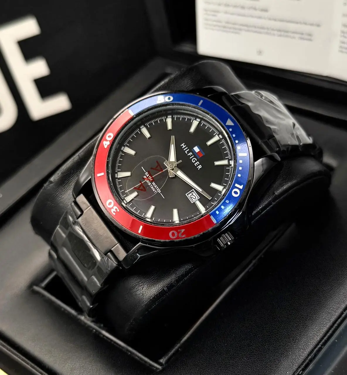 Reloj Tommy Hilfiger de acero negro para caballero con bisel rojo y azul, esfera oscura analógica, fechador y estuche acolchado.