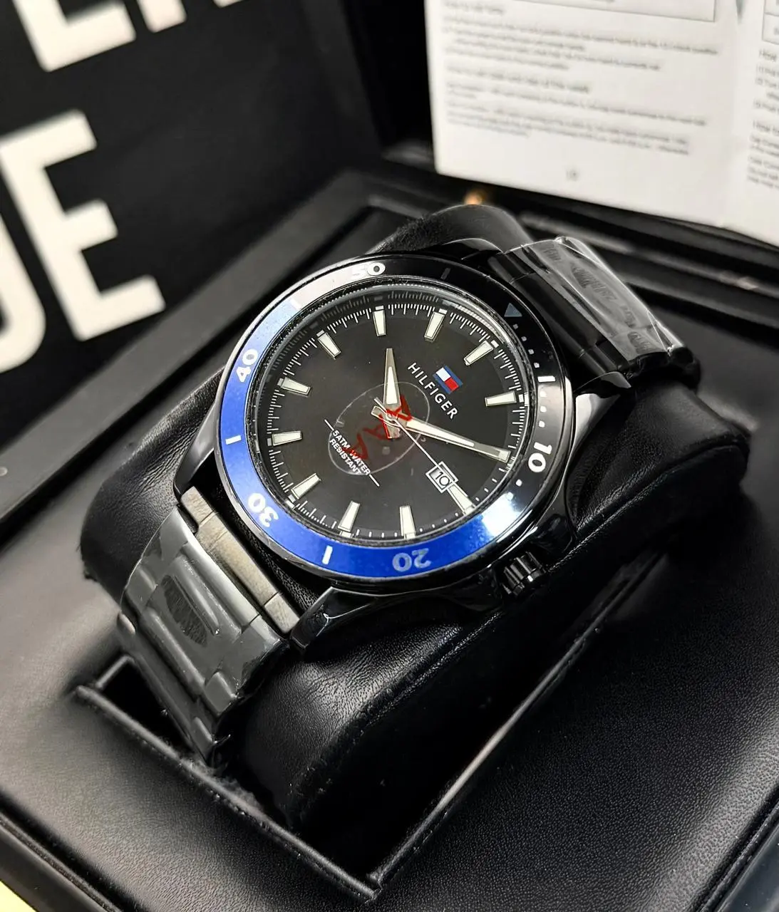 Reloj Tommy analógico de caballero con caja negra, bisel azul, esfera oscura, calendario y pulsera de acero en su estuche.