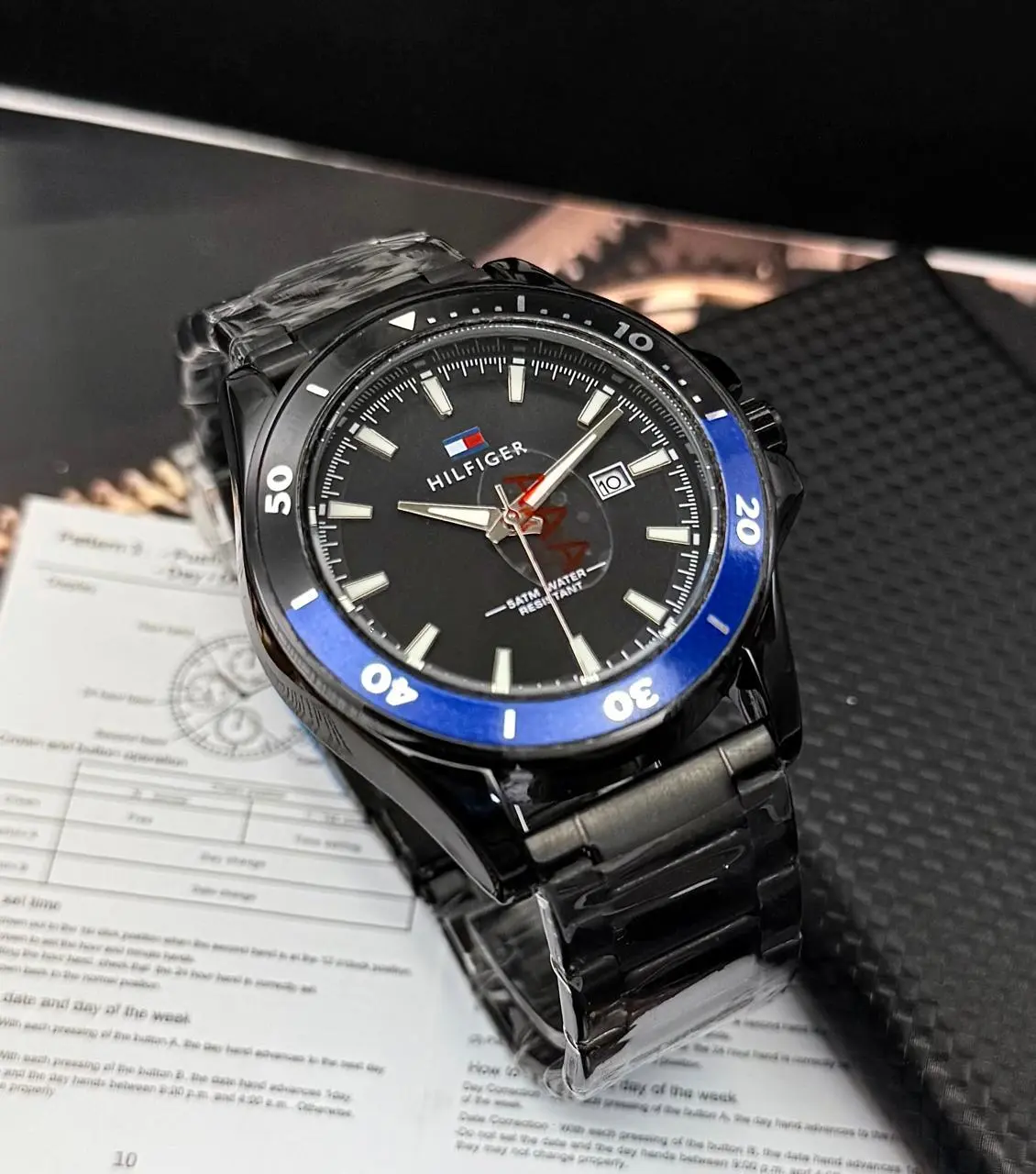 Reloj analógico Tommy para hombre de acero negro con bisel azul, fechador y fondo oscuro, sobre manual y caja.