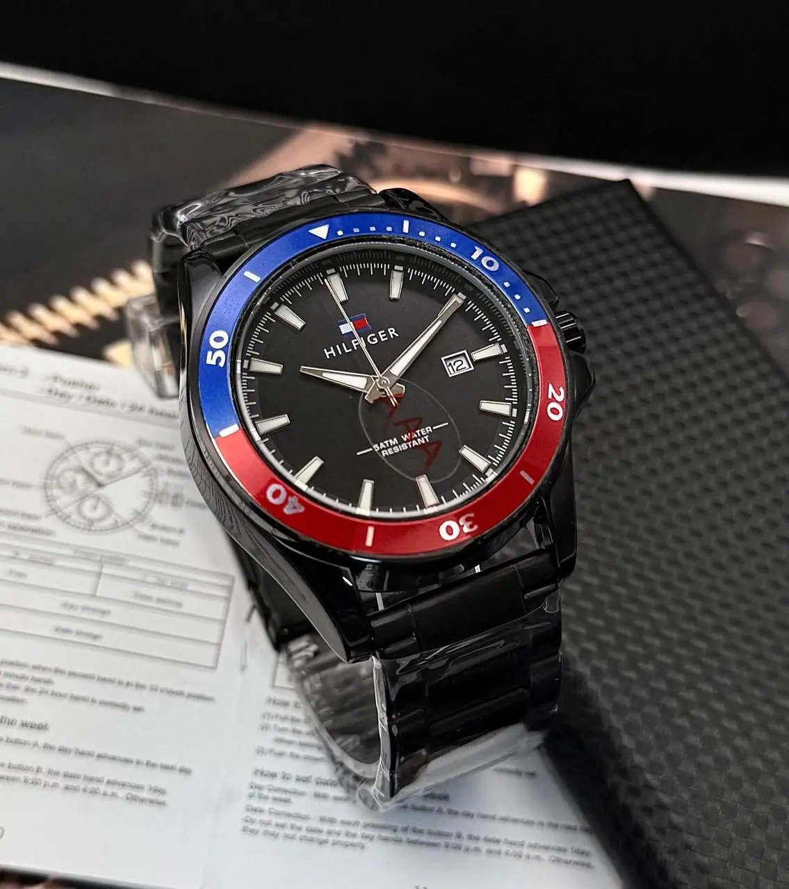Reloj Tommy analógico de acero negro para hombre con bisel giratorio azul y rojo, esfera negra, fechador y manual desplegado.