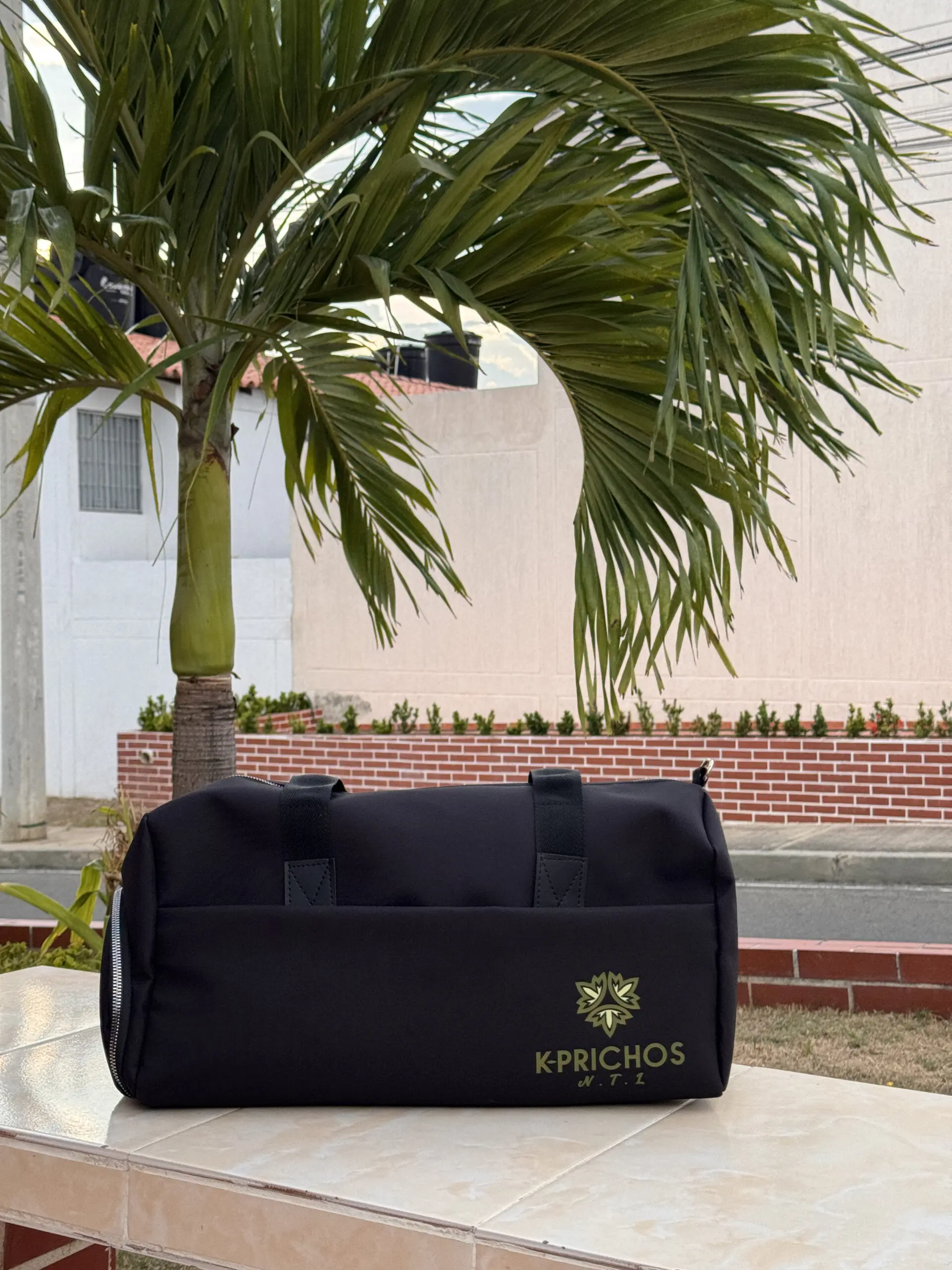 Bolso de lona negro unisex con asas cortas, logo dorado K-PRICHOS y compartimento inferior para zapatos, apoyado sobre superficie de baldosas al aire libre.