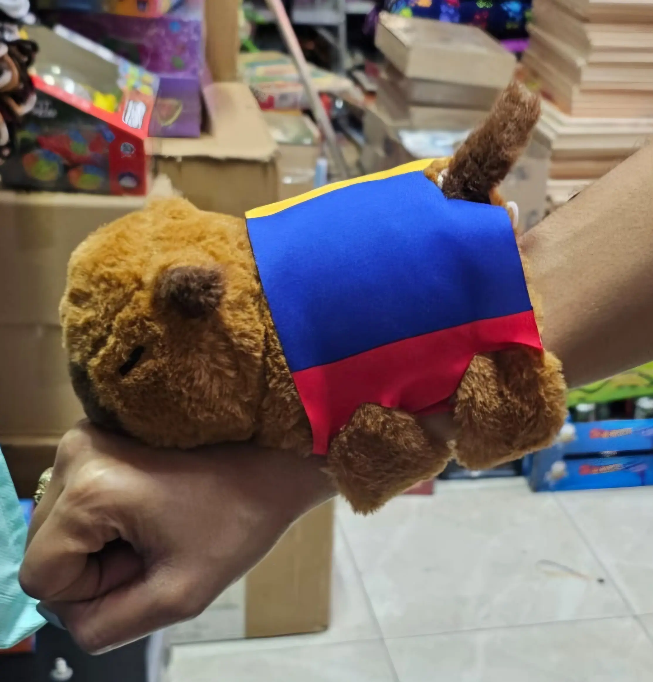 Manilla de peluche de capibara café con banda tricolor de la bandera de Colombia en amarillo, azul y rojo, sostenida por una mano.
