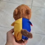 Capibara de peluche marrón con camiseta amarilla y azul, vista posterior sostenida por una mano.