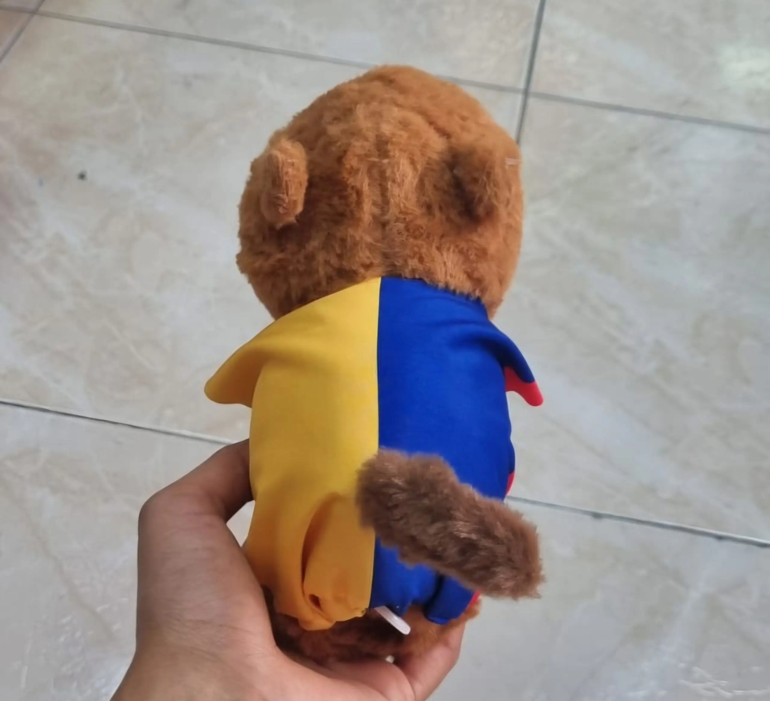 Capibara de peluche marrón con camiseta amarilla y azul, vista posterior sostenida por una mano.