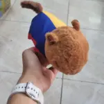 Peluche de capibara café con textura suave y prenda tricolor azul, amarilla y roja, sostenido por una mano con reloj blanco.
