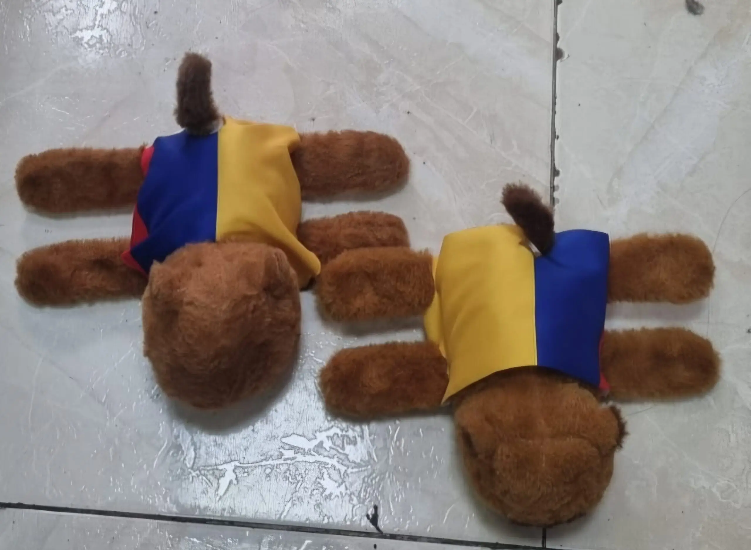 Vista superior de dos manillas de peluche de capibara marrón con bandas tricolores de Colombia sobre un piso de baldosas.