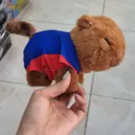 Peluche de capibara café con prenda azul y roja, sostenido por una mano para mostrar su tamaño y textura suave.