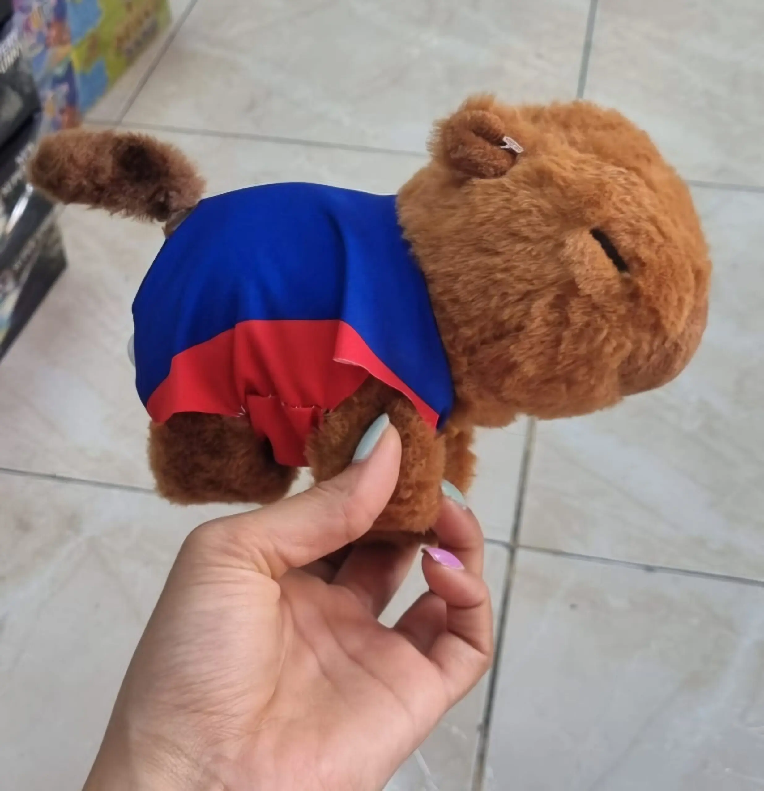 Peluche de capibara café con prenda azul y roja, sostenido por una mano para mostrar su tamaño y textura suave.