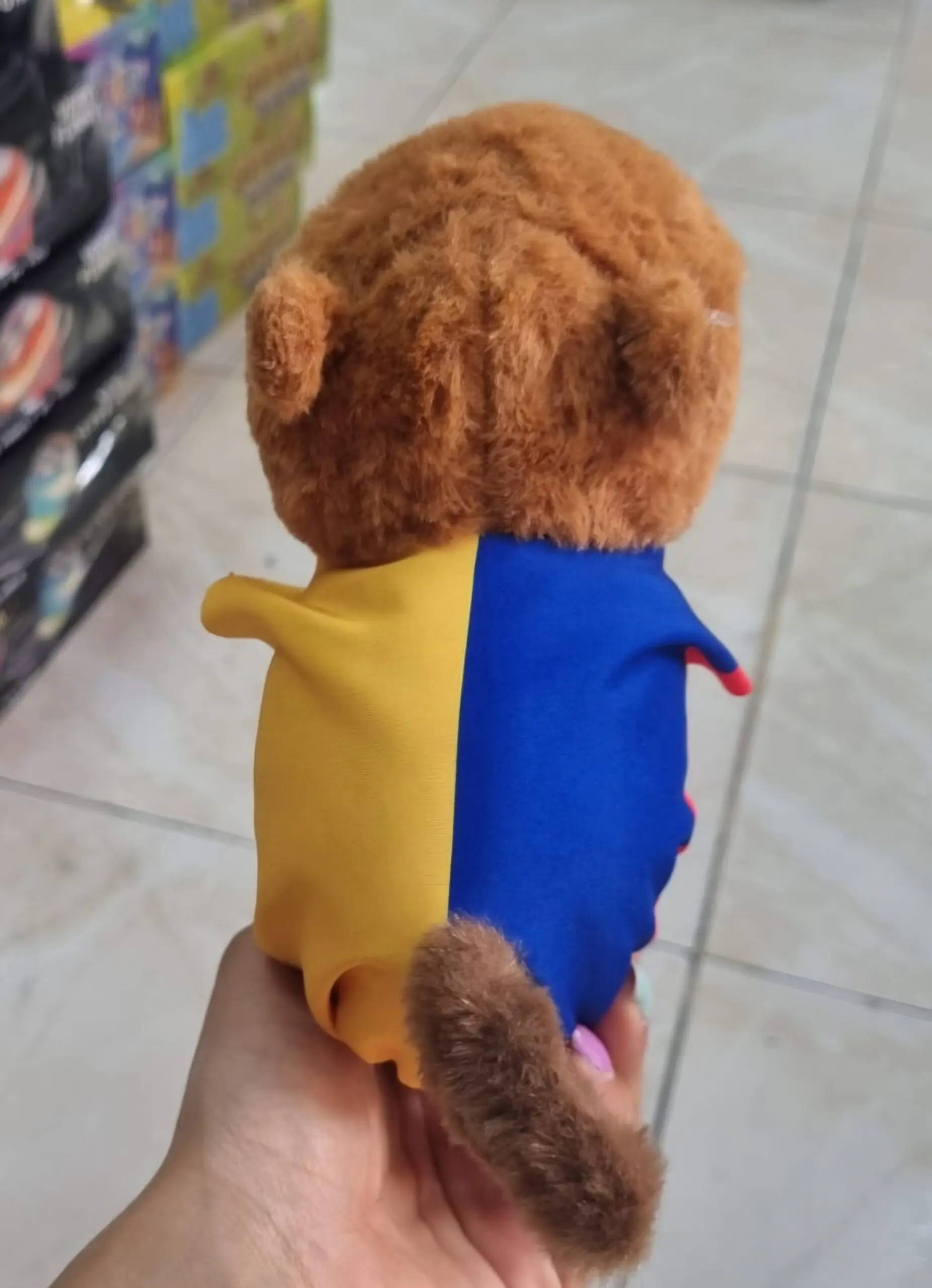 Vista posterior de peluche de capibara café con textura suave y manilla tricolor de Colombia en amarillo y azul.