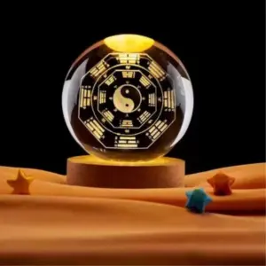 Lámpara esfera de cristal con grabado 3D Yin Yang y Bagua, luz cálida y base de madera sobre tela naranja decorativa con estrellas.