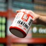 Envase blanco y rojo de Creatina Monohidrato Old School de 300g sostenido por una mano frente a estantes de suplementos.