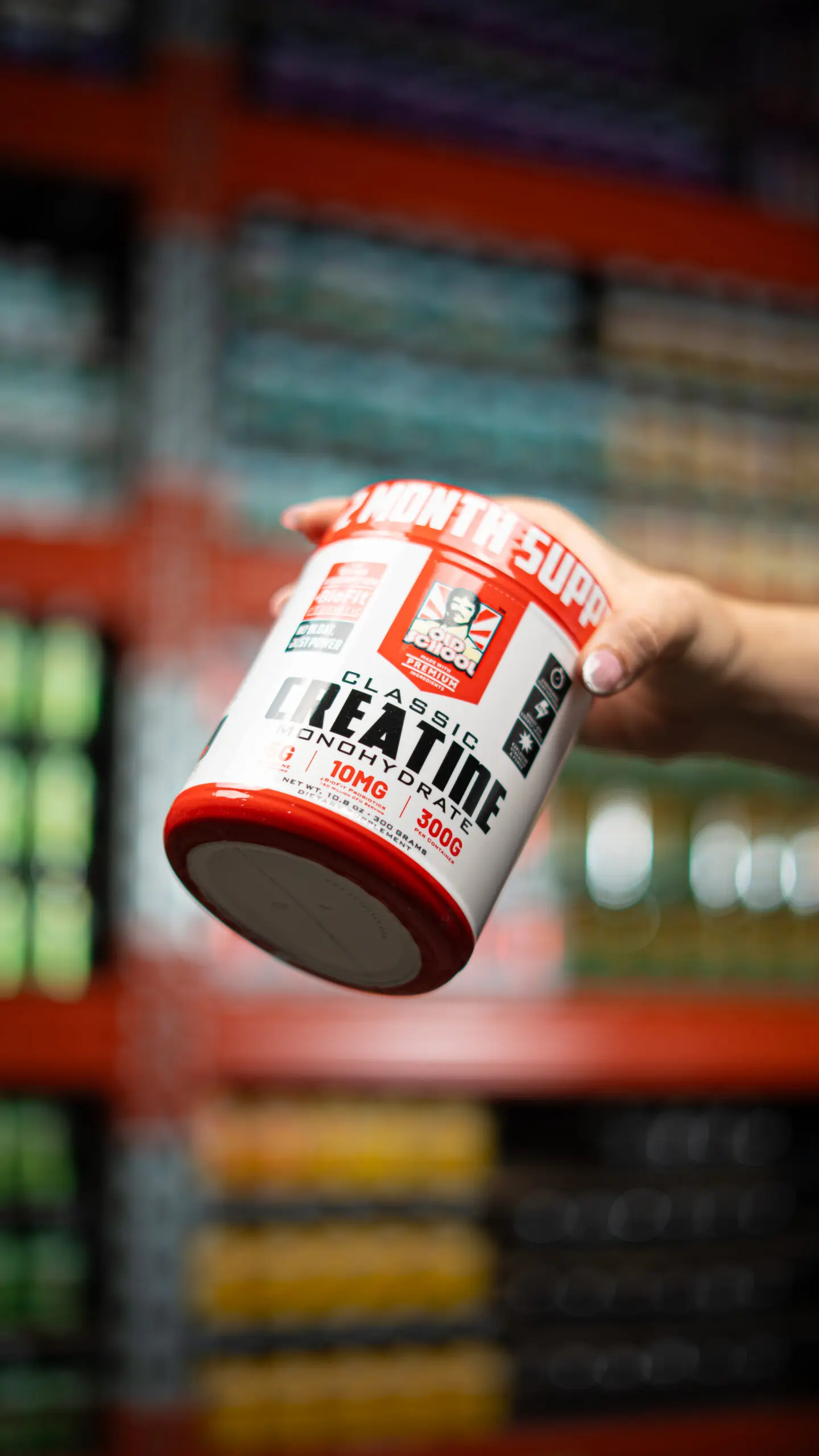 Envase blanco y rojo de Creatina Monohidrato Old School de 300g sostenido por una mano frente a estantes de suplementos.