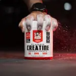 Mano con tatuaje sujeta tarro de Creatina Monohidrato Classic con polvo blanco y salpicaduras rojas sobre fondo oscuro.