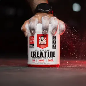 Mano con tatuaje sujeta tarro de Creatina Monohidrato Classic con polvo blanco y salpicaduras rojas sobre fondo oscuro.