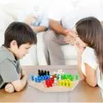 Juego de damas chinas de madera con tablero hexagonal y fichas de colores en base de madera, mientras dos niños juegan sentados alrededor de una mesa.