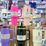 Cinco termos Stanley Quencher de 40 oz con asa y pitillo en colores rosa, morado, negro y crema, exhibidos sobre cajas en una tienda.