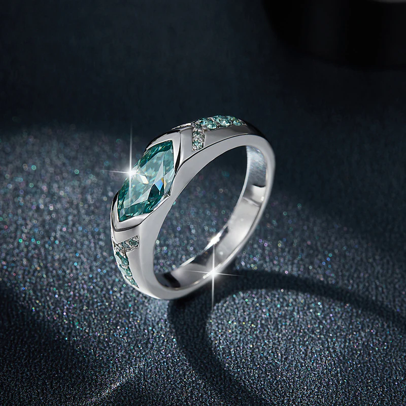Anillo de plata 925 con moissanita turquesa de corte marquise y piedras laterales, con destellos sobre fondo oscuro texturizado.