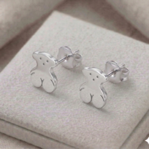 Par de aretes de broquel con forma de osito en plata 925 sobre una superficie de tela beige texturizada.