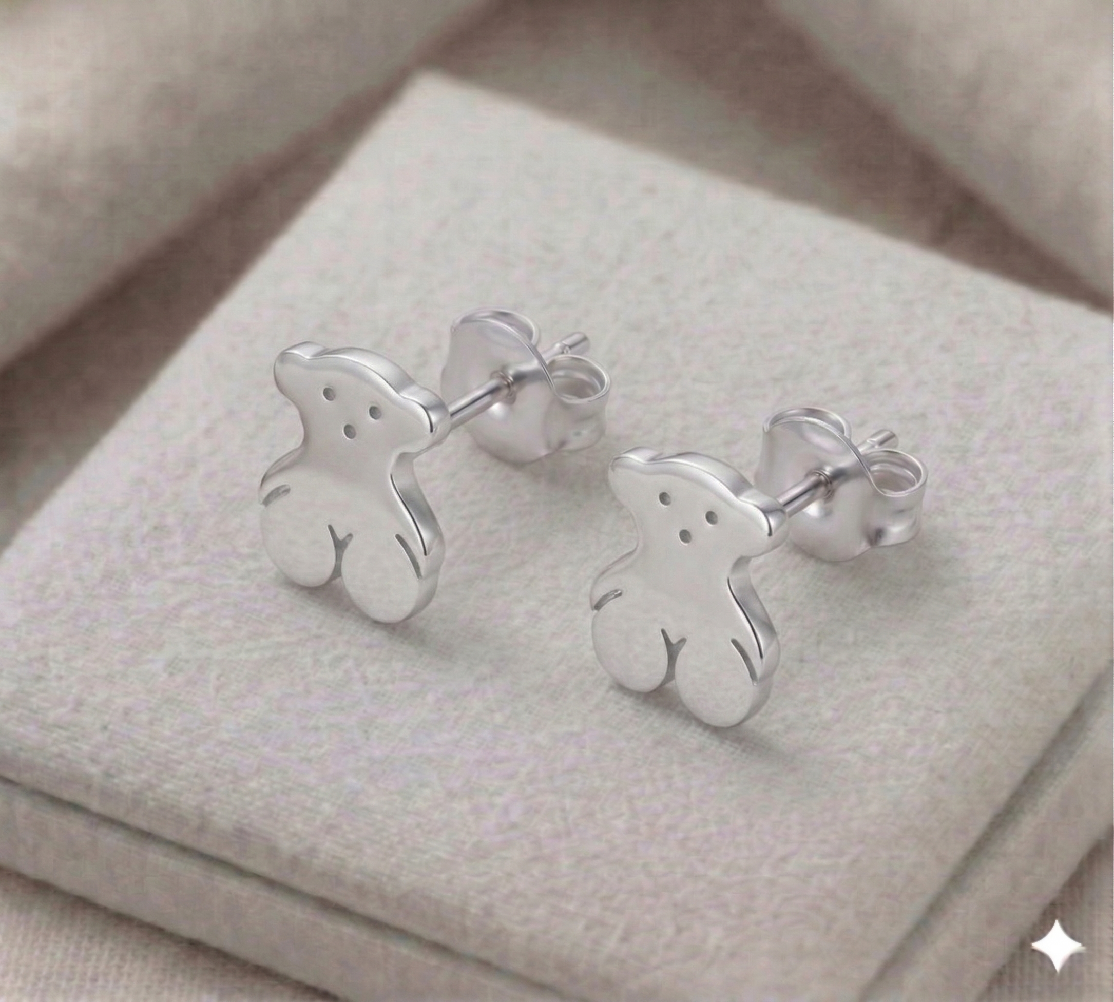 Par de aretes de broquel con forma de osito en plata 925 sobre una superficie de tela beige texturizada.