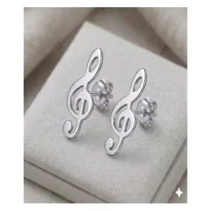 Par de aretes tipo topo en plata brillante con diseño de clave de sol, dispuestos sobre un cojín de terciopelo gris claro.
