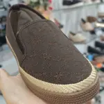 Zapato casual femenino café con bordados, ribete tejido y suela texturizada, sostenido en mano dentro de tienda.