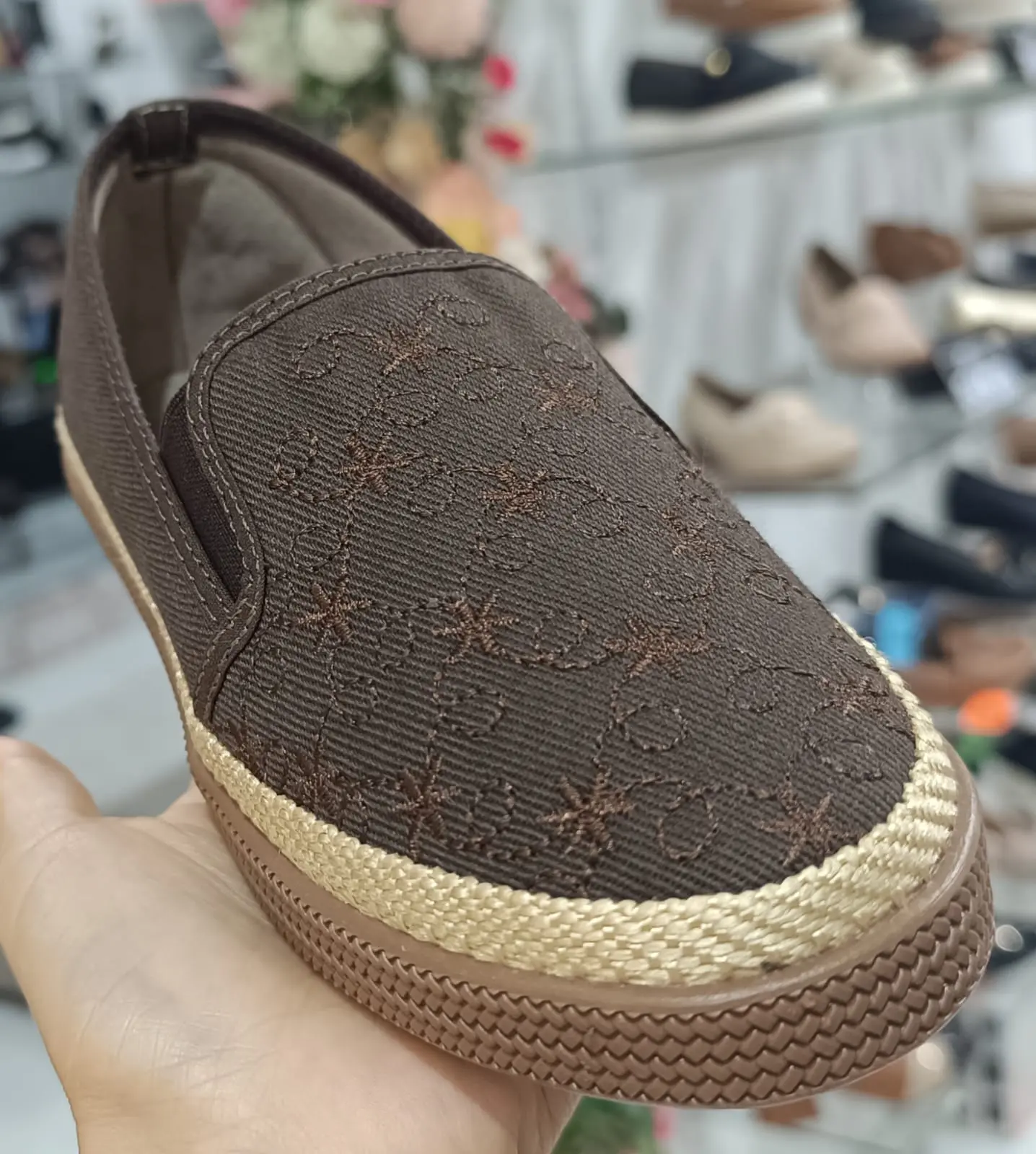 Zapato casual femenino café con bordados, ribete tejido y suela texturizada, sostenido en mano dentro de tienda.