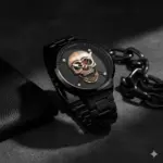 Reloj analógico negro de cuarzo con diseño de calavera dorada en relieve, sobre fondo gris con cadena metálica y billetera de cuero.